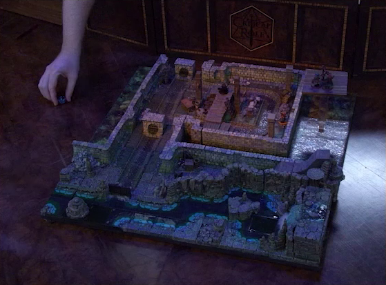 A battlemap of a stone underground room in the sewer. Brennan picks up a d20 that he just rolled off to the side of the battlemap. Bolaire, Azune, and Murray stand on a raised platform above the room indicating an upper floor, while Halandil drags a fallen Demodus Blix towards the side of the room. Crow Keepers and Laumy stand within the room. A raised wooden boardwalk goes from the wall to the center of the room among wooden boxes and three stone pillars holding up the ceiling. An underground river runs past the room on the south end.
