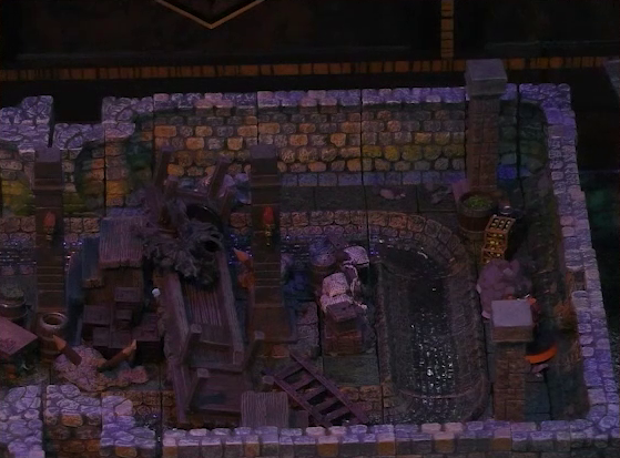A battlemap of a stone underground room in the sewer. A wooden boardwalk is collapsed in the center of the room, with Laumy standing atop the wreckage. Halandil stands by the wall of the room. A sewer trench runs in a circle in the room around pillars holding up the ceiling.