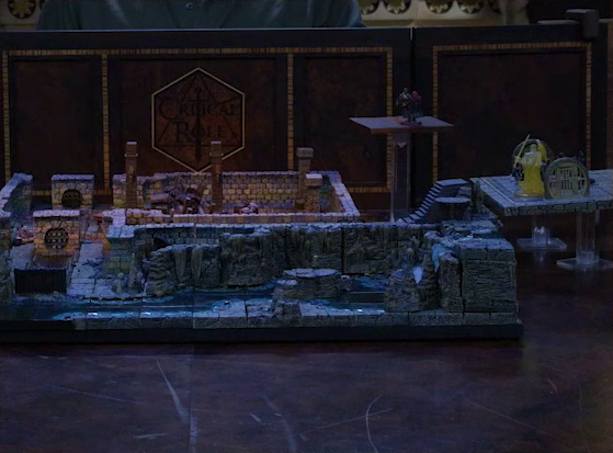A battlemap of an underground area in the sewer. Two raised platforms indicating multiple levels rise above a stone room. Bolaire and Halandil stand on the higher one, while a knight made of light, Azune, Murray, Demodus Blix, and four Crow Keepers are on the lower one. The room has sewer grates at north and south entrances. A river runs past the room, and past a hallway with stone stairs leading down into it.