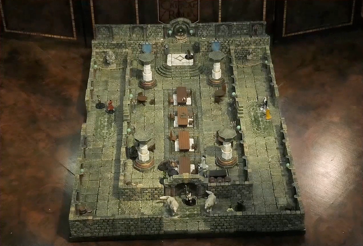 A battlemap of a large stone banquet chamber with surrounding hallways. The chamber has four entrances, one on each side. At the south entrance is Filoneus Halovar with his two dogs Wrath and Judgement. Inside the chamber, King Augustus Valian, a Hound, and a druid sit across a table from Photarch Yanessa and an attendant Lux. Murray hides invisibly behind a pillar near the table. Another Hound stands at the north entrance. Azune and Bolaire are at the end of the north hallway. Six guests amble around the surrounding hallways.