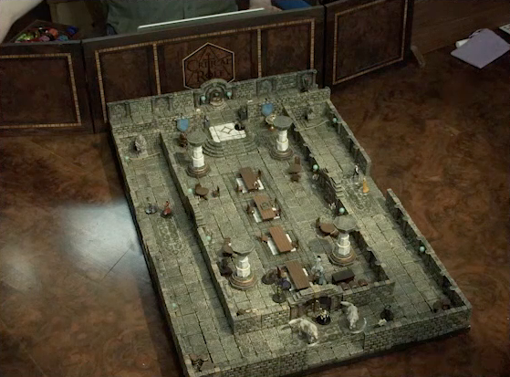 A battlemap of a large stone banquet chamber with surrounding hallways. The chamber has four entrances, one on each side. At the south entrance of the chamber is Filoneus Halovar with his two dogs Wrath and Judgement. Inside the chamber, King Augustus Valian, a Hound, and a druid sit across a table from Photarch Yanessa and an attendant Lux. Murray hides invisibly behind a pillar near the table. Another Hound stands at the north entrance. Azune and Bolaire are at the end of the north hallway. Six guests amble around the surrounding hallways. Next to the map is a DM screen, behind which Brennan stands.