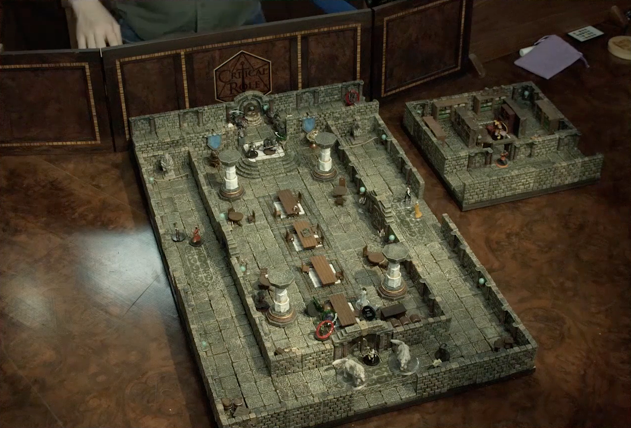 A small battlemap of a library chamber, placed on the table next to a battlemap of a large stone banquet chamber with surrounding hallways. Halandil stands in the open doorway to the library, looking in at a Nama. The library is filled with bookshelves. In the larger battlemap, King Augustus Valian and a Hound, both with green “POISONED” status rings around them, are on one side of a table. A druid next to them has a red ring. On the other side of the table Photarch Yanessa lays on the ground next to an attendant Lux. Murray invisibly hides behind a nearby pillar. Outside the south entrance of the chamber are Filoneus Halovar with his two dogs Wrath and Judgement. Azune and Bolaire are at the end of the north hallway. Halfway down that hallway six assassins burst past a Hound of the King to enter the chamber’s north entrance.