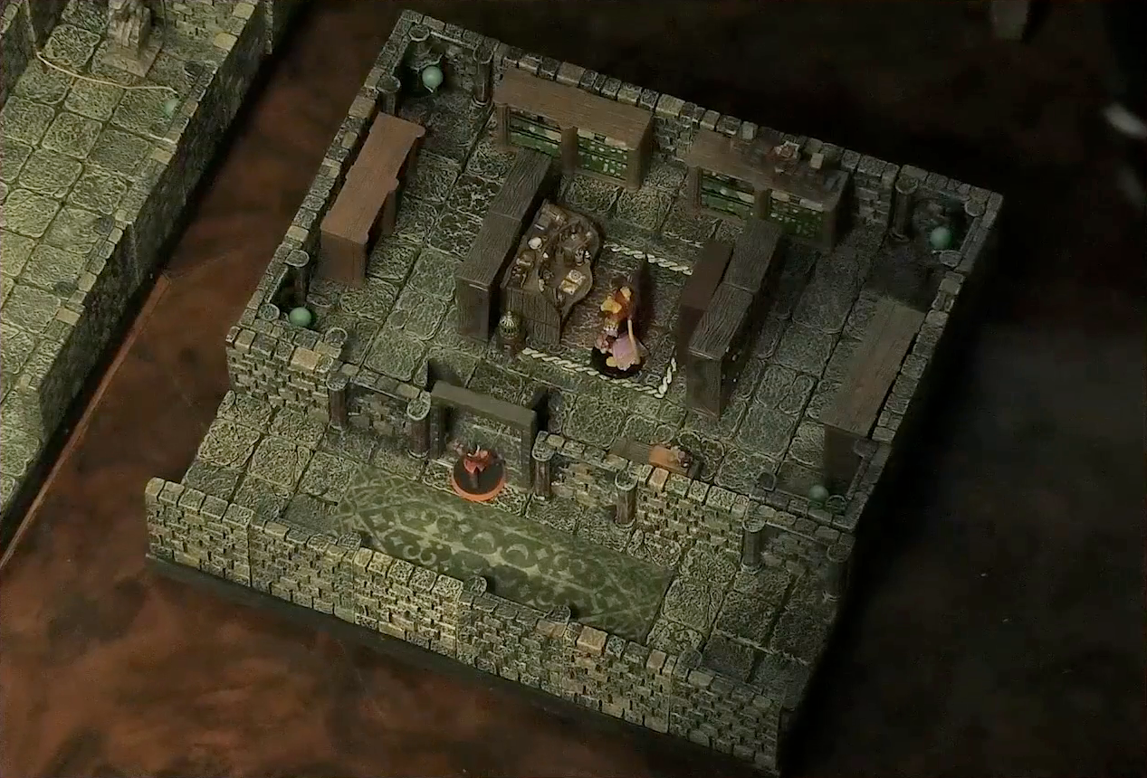 A small battlemap of a library chamber. Halandil stands in the open doorway to the library, looking in at a Nama. The library is filled with bookshelves and one desk cluttered with books and trinkets. The Nama stands next to the desk. Next to the battlemap at the edge of the image is a larger battlemap.