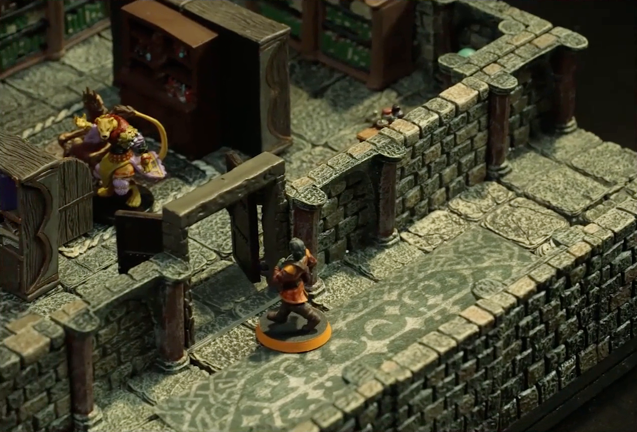 A small battlemap of a library chamber. Halandil stands in the open doorway to the library, looking in at a Nama. The library is filled with bookshelves and one desk cluttered with books and trinkets. The Nama stands next to the desk.