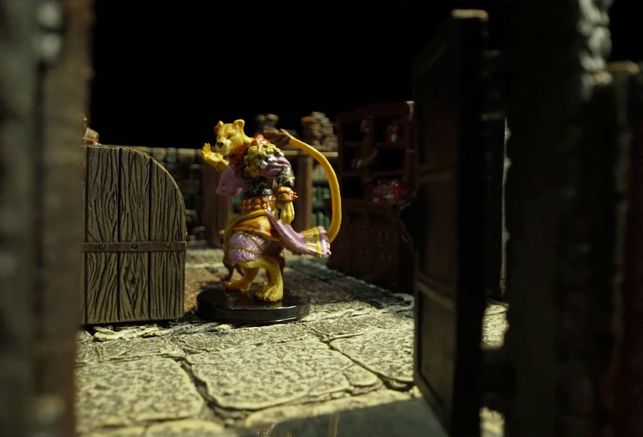 A small battlemap of a library chamber focused on a yellow Nama, viewed through an open doorway. The Nama wears a mixture of fabrics in colors red, yellow, green, and purple. His tail curls back behind its head.