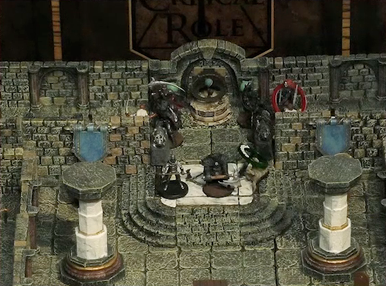 A battlemap of a large stone banquet chamber with surrounding hallways. Azune and Bolaire face off against six assassins entering the chamber through its north entrance. A fallen Hound of the King with a “POISONED” status ring lays on the ground just inside the entrance. Bolaire has a red status ring around his miniature. Inside the chamber are marble pillars holding up the ceiling.