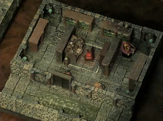 A small battlemap of a library chamber. Halandil stands just inside its closed door. A Nama with a “CONCENTRATION” status ring around its miniature stands in the corner of the room, a book shelf in between him and Halandil. In the center of the room among the book shelves is a desk cluttered with books and trinkets. In front of the desk is a red padded chair.
