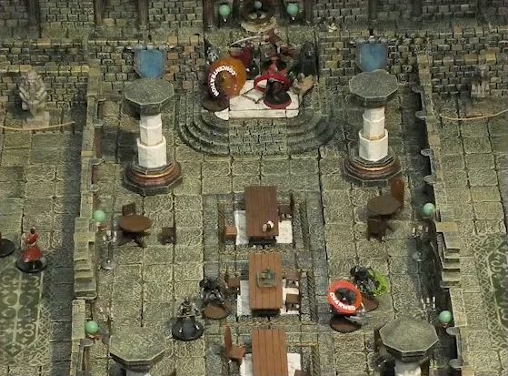 A battlemap of a large stone banquet chamber with surrounding hallways. At the north entrance, Azune and Bolaire fight two assassins. Bolaire has a red status ring around his miniature, and one of the assassins has a “CONCENTRATION” status ring around it. A Flaming Sphere is amidst the melee. Halfway into the room are four more assassins, one with a “CHARMED” ring and another with a green ring. Two party guests are in the west hallway outside the chamber.