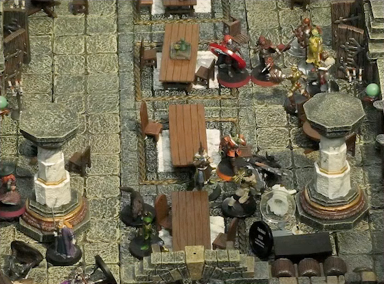 A battlemap of a large stone banquet chamber with surrounding hallways. There is a chaotic scene, where Filoneus Halovar stands over the body of the fallen Photarch Yanessa, as well as a fallen assassin. Halandil stands right next to them, as does a Timmonish knight. Bolaire, Azune, and a number of guards approach from the north. Murray, King Augustus Valian, a Hound, and a druid stand on the opposite side of the room near a pillar. A fallen attendant Lux lies next to the Photarch, and two fallen assassins are next to the King.