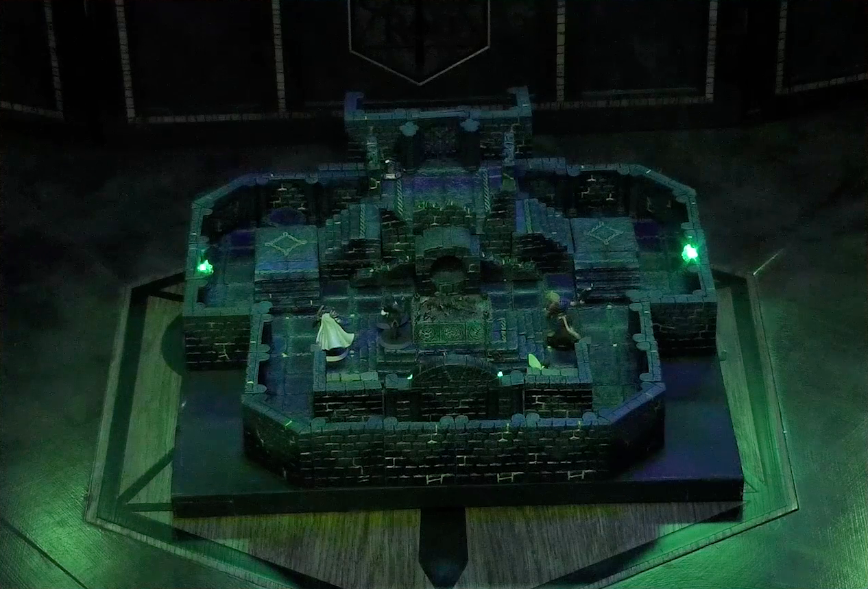 A battlemap of a stone chamber on the Critical Role table. A stone altar is in the center with a skeletal corpse on top of it, next to which stand Occtis and Pincushion. Around the altar are Thimble, Julien, Teor, and Talcydimir. Behind the altar is a circular hole leading into the wall. Two staircases on either side of the hole lead up to a door with a portcullis. The room is lit by a sickly green glow emanating from torches on the walls.