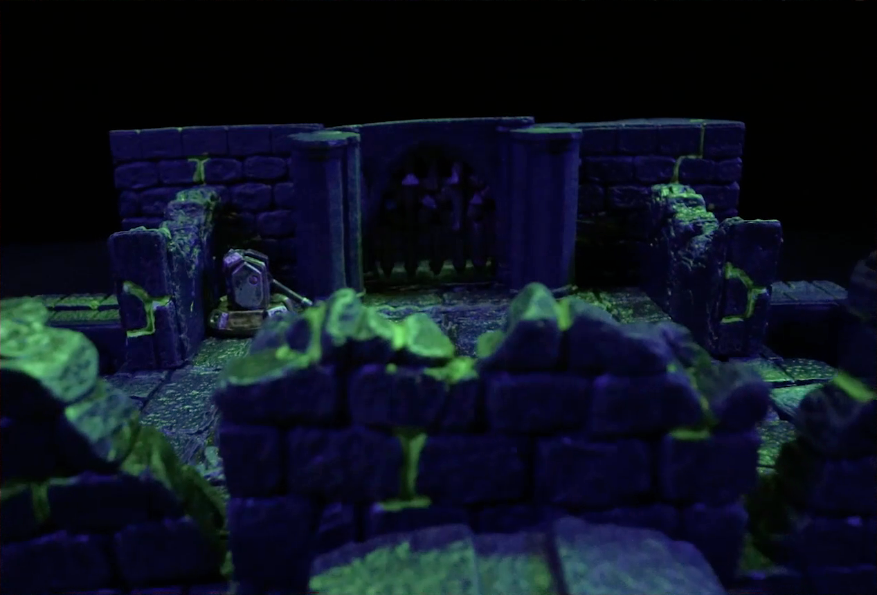 A battlemap of a stone chamber. A portcullis sits closed next to a lever which is in the down position. The camera views the portcullis from behind a stone railing. The room is lit by a sickly green glow.