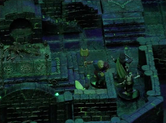 A battlemap of a stone chamber. Talcydimir faces off against an armored skeleton which is standing behind Julien. Thimble is around a corner from the skeleton. Off to their side is a stone altar with a skeletal corpse on top of it. Behind the altar is a circular hole leading into the wall. A staircase rises up to the side of the hole. The room is lit by a sickly green glow emanating from torches on the walls.