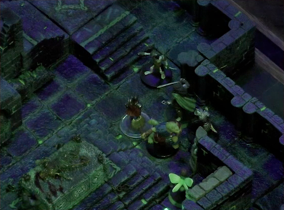 A battlemap of a stone chamber. Talcydimir, Dame Gaya Seremai, Julien, and Thimble face off against two armored skeletons. Off to their side is a stone altar with a skeletal corpse on top of it. Behind the altar is a circular hole leading into the wall. A staircase rises up to the side of the hole. The room is lit by a sickly green glow.