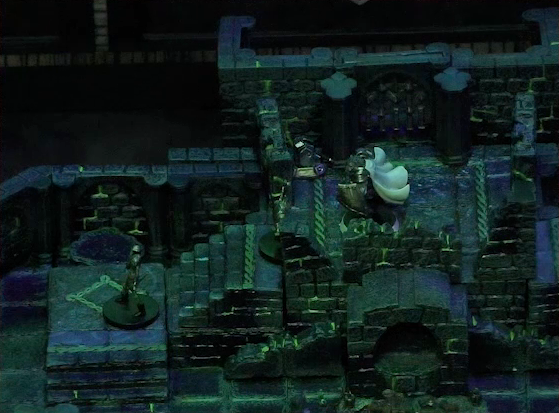 A battlemap of a stone chamber. Teor stands at the top of a staircase with two armored skeletons climbing the stairs towards him. To Teor’s right is a door blocked by a portcullis, next to which is a lever in the down position. In front of the stairs is a circular hole leading into the wall. The room is lit by a sickly green glow.