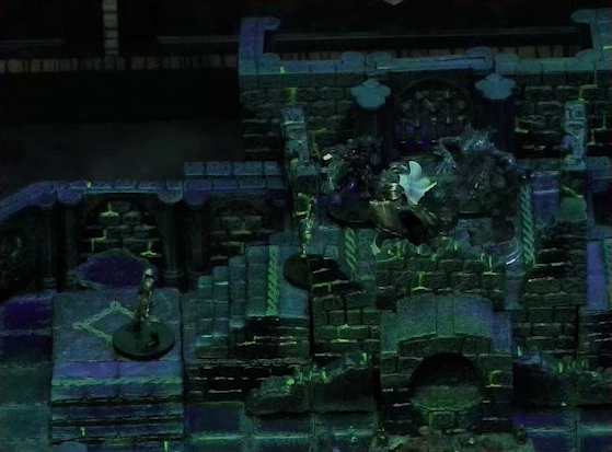 A battlemap of a stone chamber. Teor stands at the top of a staircase surrounded by four shadows, with two armored skeletons climbing the stairs towards him. To Teor’s right is a door blocked by a portcullis, next to which is a lever in the down position. In front of the stairs is a circular hole leading into the wall. The room is lit by a sickly green glow.