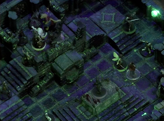 A battlemap of a stone chamber. Teor and Talcydimir stand at the top of a staircase surrounded by four shadows, with two armored skeletons climbing the stairs towards them. To their right is a door blocked by a portcullis, next to which is a lever in the down position. In front of the stairs is a circular hole leading into the wall. In front of the hole is a stone altar, on which stands Occtis. Thimble, Dame Gaya Seremai, and Julien face off against an armored skeleton near the altar, Julien standing on the base of the stairs. Pincushion stands on the opposite side of the altar from them. The room is lit by a sickly green glow emanating from torches on the walls.