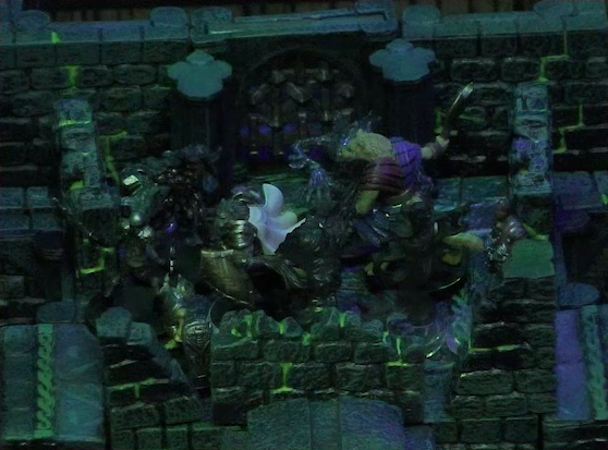 A battlemap of a stone chamber. Teor and Talcydimir stand at the top of a staircase surrounded by four shadows. Dame Gaya Seremai helps them from the other side of the shadows. To their right is a door blocked by a portcullis, next to which is a lever in the down position. In front of the stairs is a circular hole leading into the wall. The room is lit by a sickly green glow.