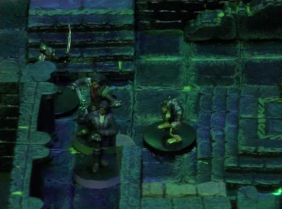 A battlemap of a stone chamber. Julien and Occtis face off against two armored skeletons. One is short and stocky, the other wields a long curved blade. Julien and Occtis stand against the wall, with short stone steps leading up to an altar just out-of-frame on their other side. The room is lit by a sickly green glow.