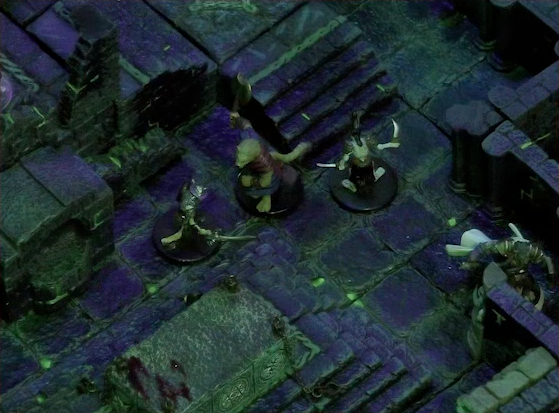 A battlemap of a stone chamber. Talcydimir faces off against two armored skeletons, while Teor faces away heading down a hallway. Off to Talcydimir’s side is a stone altar. Behind the altar is a circular hole leading into the wall. A staircase rises up to the side of the hole. The room is lit by a sickly green glow.