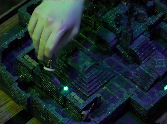 A battlemap of a stone chamber. Brennan’s hand reaches in to lift Teor out of the map near a revealed secret door in a side wall. Talcydimir stands near the door as well. A stone altar is in the center of the map. Behind the altar is a circular hole leading into the wall. Two staircases on either side of the hole lead up to a door with a portcullis, in front of which stands a group of shadows. The room is lit by a sickly green glow emanating from torches on the walls.