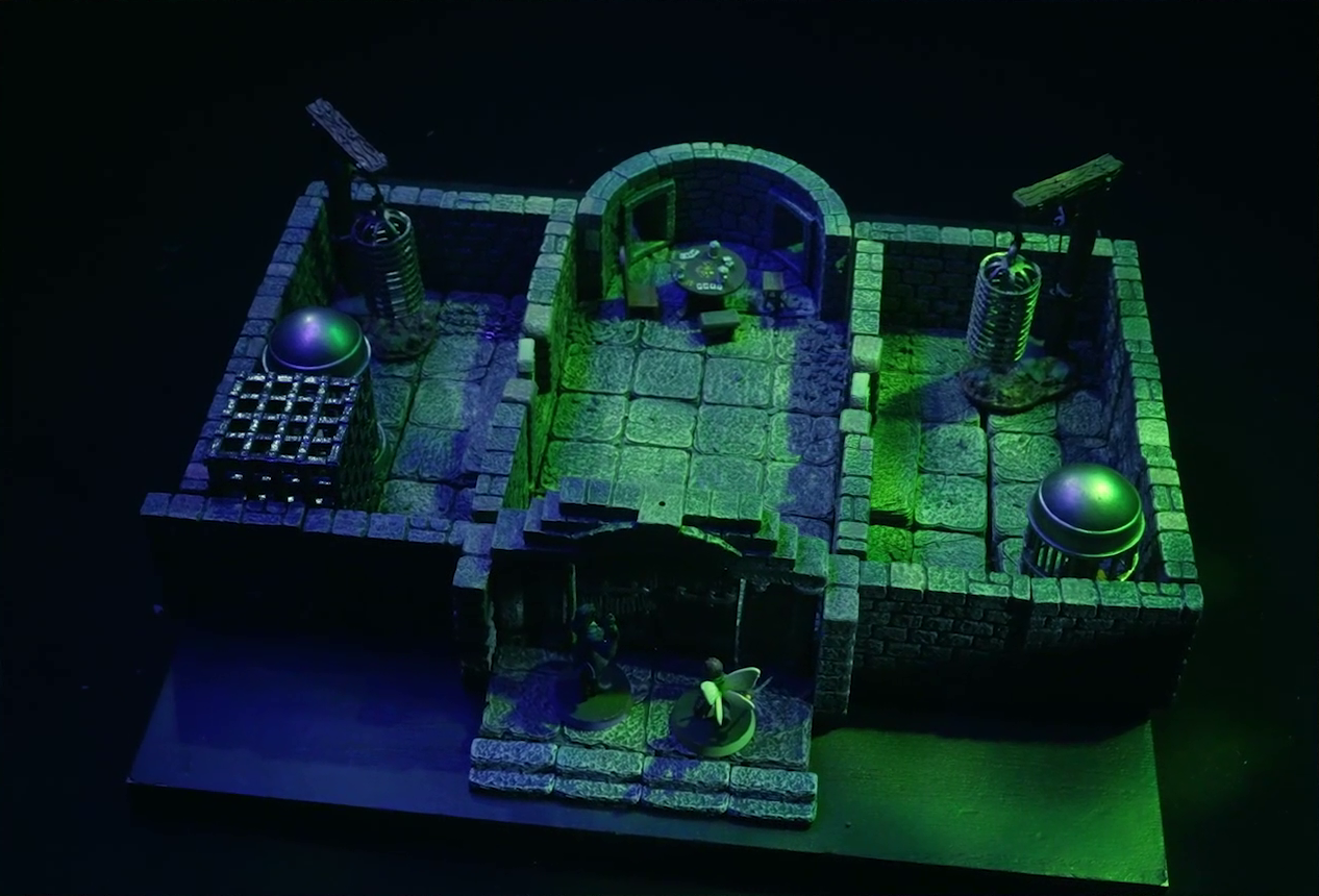 A battlemap of a stone aviary split into three rooms. Occtis and Thimble stand outside a closed door into the main chamber, in which is a circular table with a card game abandoned on it. To the left of the main chamber is a closed door leading to a room with a large cubic black metal cage, a large cylindrical silvery metal cage, and a large cylindrical metal hanging cage resembling a bird cage. To the right of the main chamber is a closed door leading to a room with the same kind of silvery metal cage and hanging cage, although there is no large cubic cage.