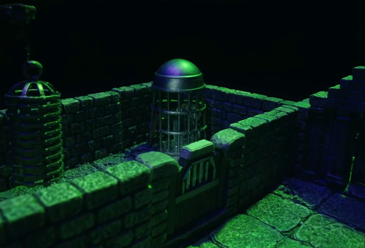A battlemap of a stone aviary focused on a large cylindrical silvery metal cage in a room. Inside the cage is a flying brown owl with barely any room to move around. Next to this cage is a large cylindrical metal hanging cage resembling a bird cage, which is empty. The cages are in a small room with its one door shut. Outside the room is another stone chamber with a closed wooden door on one wall.