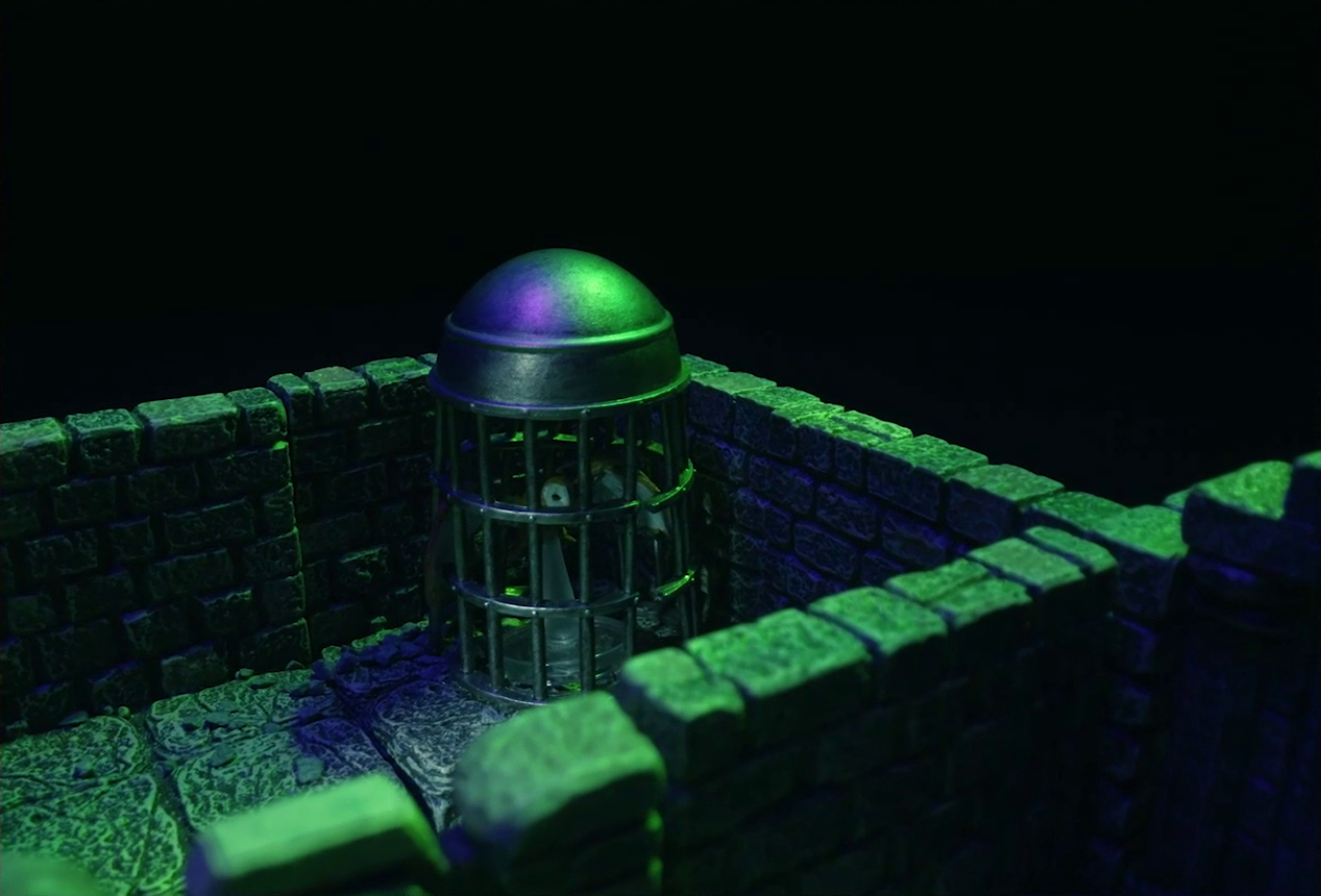 A battlemap of a stone aviary focused on a large cylindrical silvery metal cage in a room. Inside the cage is a flying brown owl with barely any room to move around. The owl’s wings are outspread, its right wing poking out slightly through the bars. The owl’s face and the underside of its wings are white. Its eyes are black. The cage is in a small room on the other side of a wall from another room, only the corner of which is visible.