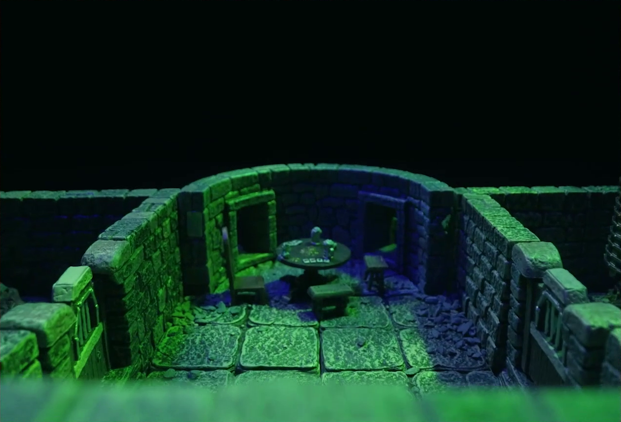 A battlemap of a stone aviary focused on a main chamber with two other rooms on either side of it. At the rear end of the chamber is a circular wooden table with a card game abandoned on it. A couple drinking vessels sit next to the cards. Around the table are a wooden chair and two short wooden stools. The two rooms off the main chamber have their wooden doors closed. In the right room, the edge of a large cylindrical metal cage is just visible.