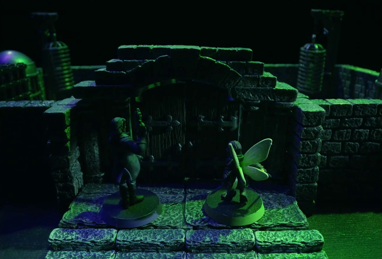 A battlemap focused on Occtis and Thimble standing outside the closed wooden double doors to a stone aviary. Off to either side of the main chamber of the aviary are two rooms. The left room has a large cubic black metal cage, a large cylindrical silvery metal cage, and a large cylindrical metal hanging cage resembling a bird cage. The right room has the same kind of silvery metal cage and hanging cage, although there is no large cubic cage.