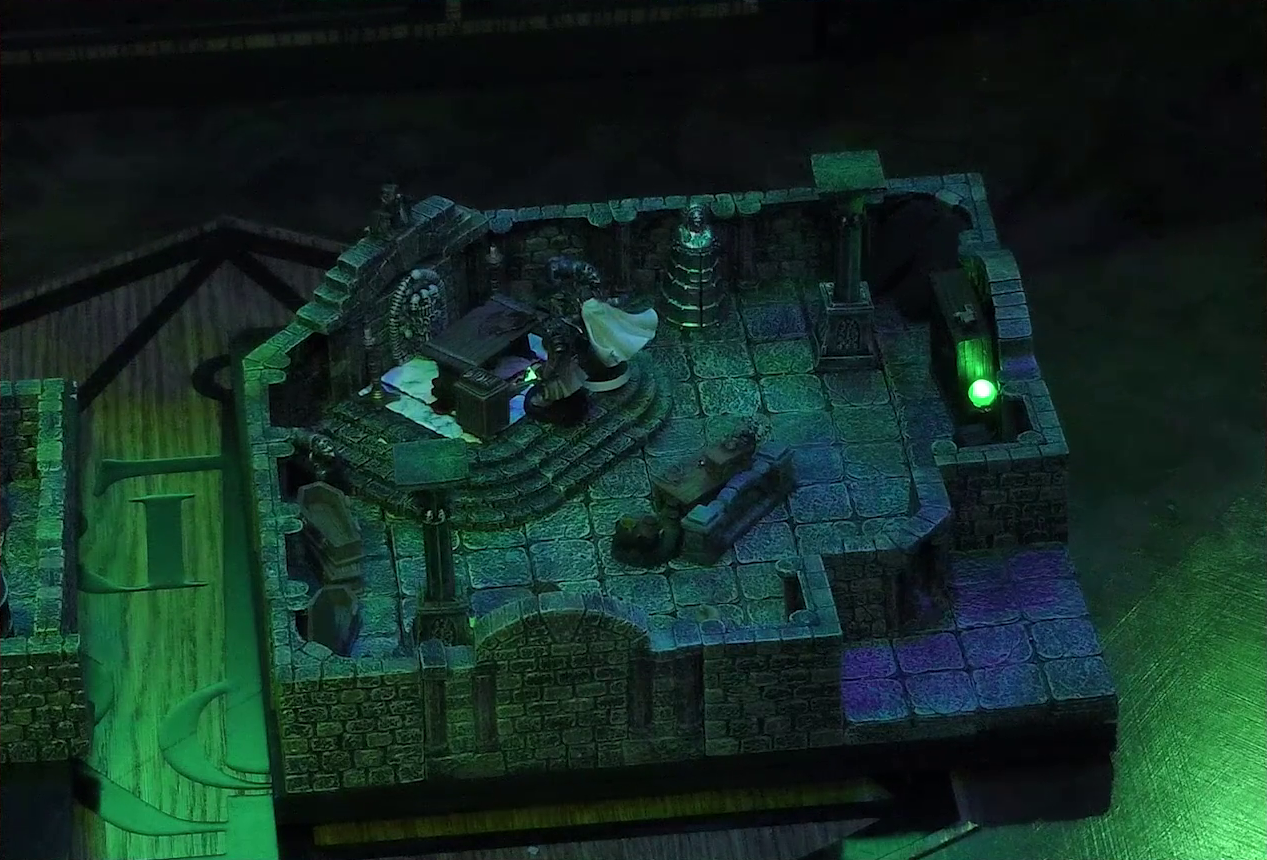 A battlemap of a stone crypt with open wooden coffins on one wall and a silvery metal iron maiden on another wall. Teor and Talcydimir stand in front of a wooden table with a splatter of blood on top of it. The table sits on a raised part of the floor in front of a wall decorated with carved skull motifs. On a box in front of the table sits a tray bearing multiple surgical instruments. The room is lit by eerie glowing green torches.
