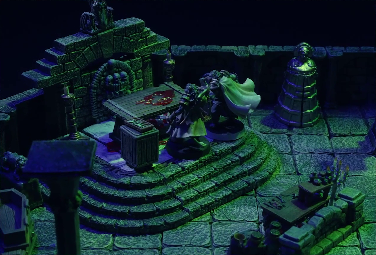 A battlemap of a stone crypt with open wooden coffins on one wall and a silvery metal iron maiden on another wall. Teor and Talcydimir stand in front of a wooden table with a splatter of blood on top of it. The table sits on a raised part of the floor in front of a wall decorated with carved skull motifs. On a box in front of the table sits a tray bearing multiple surgical instruments. The room is lit by eerie green light.