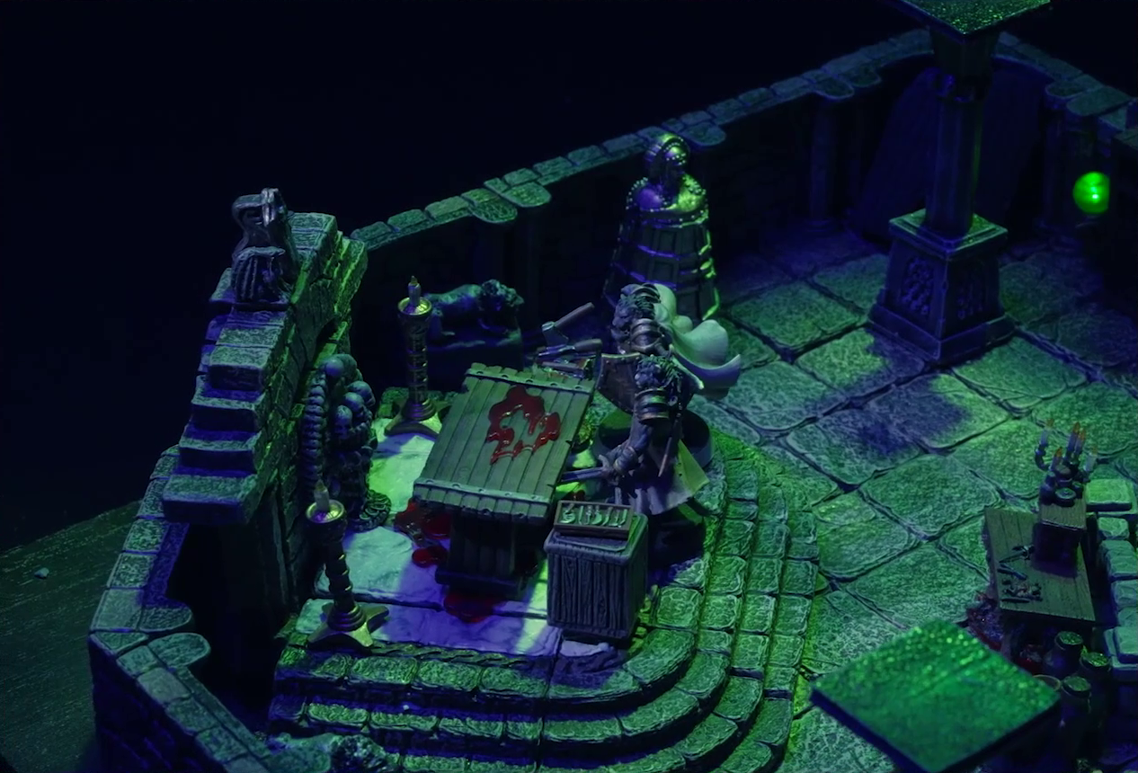 A battlemap of a stone crypt with a silvery metal iron maiden on one wall. Teor and Talcydimir stand in front of a wooden table with a splatter of blood on top of it. The table sits on a raised part of the floor in front of a wall decorated with carved skull motifs. On a box in front of the table sits a tray bearing multiple surgical instruments. The room is lit by eerie glowing green torches.