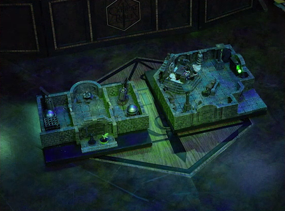 Two battlemaps placed side-by-side on the Critical Role table. The left battlemap is a stone aviary split into three rooms. Occtis and Thimble stand outside the closed door into the aviary’s main chamber. In the two side rooms are a number of large cages. The right battlemap is a stone crypt. Teor and Talcydimir stand in front of a wooden table with a splatter of blood on top of it. The table sits on a raised part of the floor in front of a wall decorated with carved skull motifs.