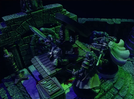 A battlemap of a stone crypt focused on Teor and Talcydimir standing in front of a wooden table on which an undead Raimond Davinos is lying. Raimond is in full armor and wielding a large double-edged sword. Behind the table is a wall decorated with carved skull motifs. On a box in front of the table sits a tray bearing multiple surgical instruments. The room is lit by eerie green light.