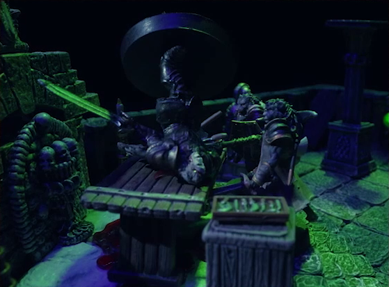 A battlemap of a stone crypt focused on Teor and Talcydimir standing in front of a wooden table on which an undead Raimond Davinos is lying. Raimond is in full armor and wielding a large double-edged sword. Behind the table is a wall decorated with carved skull motifs. On a box in front of the table sits a tray bearing multiple surgical instruments. The room is lit by eerie green light.