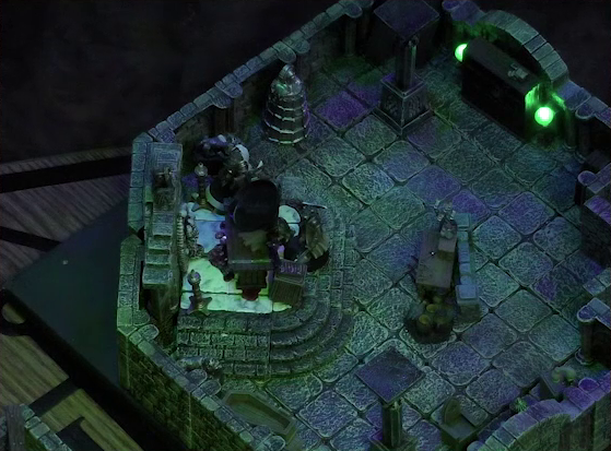A battlemap of a stone crypt with open wooden coffins on one wall and a silvery metal iron maiden on another wall. Teor and Talcydimir stand next to a wooden table on which an undead Raimond Davinos is lying. Teor is positioned on the opposite side of the table from Talcydimir, flanking the prone Raimond. The table sits on a raised part of the floor in front of a wall decorated with carved skull motifs. On a box in front of the table sits a tray bearing multiple surgical instruments. The room is lit by eerie glowing green torches.