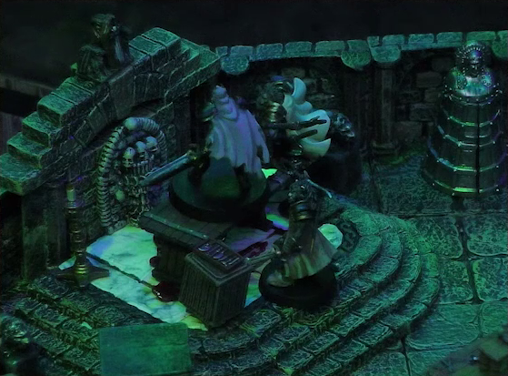 A battlemap of a stone crypt focused on Teor and Talcydimir standing next to a wooden table on which an undead Raimond Davinos is standing. Teor is positioned on the opposite side of the table from Talcydimir, flanking Raimond. The table sits on a raised part of the floor in front of a wall decorated with carved skull motifs. On a box in front of the table sits a tray bearing multiple surgical instruments. A silvery metal iron maiden is against a nearby wall. The room is lit by eerie green light.