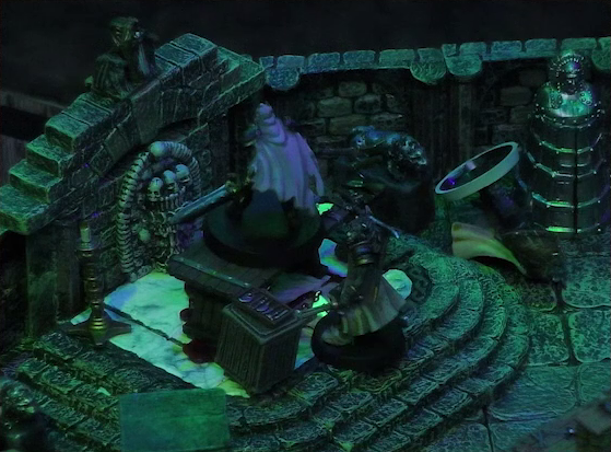 A battlemap of a stone crypt focused on a wooden table on which an undead Raimond Davinos is standing. Teor lies on the ground a few feet from the table, while Talcydimir stands nearby facing Raimond. The table sits on a raised part of the floor in front of a wall decorated with carved skull motifs. On a box in front of the table sits a tray bearing multiple surgical instruments. A silvery metal iron maiden is against a nearby wall. The room is lit by eerie green light.