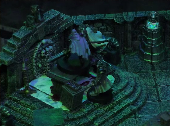A battlemap of a stone crypt focused on Teor and Talcydimir standing next to a wooden table on which an undead Raimond Davinos is standing. Teor is positioned on the opposite side of the table from Talcydimir, flanking Raimond. The table sits on a raised part of the floor in front of a wall decorated with carved skull motifs. On a box in front of the table sits a tray bearing multiple surgical instruments. A silvery metal iron maiden is against a nearby wall. The room is lit by eerie green light.