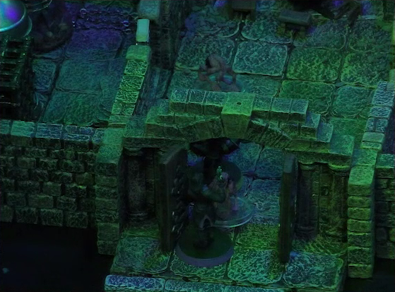 A battlemap of a stone aviary focused on Occtis, standing in the open door to a main chamber, facing off against a ghoul. Further into the chamber Occtis’s undead monitor lizard faces another ghoul. To the left of the chamber is a closed door leading to a room with a large cubic black metal cage, a large cylindrical silvery metal cage, and a large cylindrical metal hanging cage resembling a bird cage.