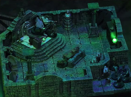A battlemap of a stone crypt. Teor and Talcydimir stand next to a wooden table on which an undead Raimond Davinos is standing. A hoard of eight zombies enters the crypt from across the room from the table. Two are in the room, the other six are just outside. Teor is positioned on the opposite side of the table from Talcydimir, flanking Raimond. The table sits on a raised part of the floor in front of a wall decorated with carved skull motifs. On a box in front of the table sits a tray bearing multiple surgical instruments. The room is lit by eerie glowing green torches.