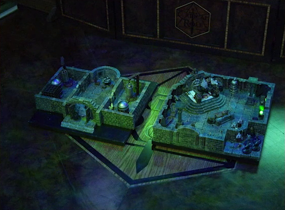 Two battlemaps placed side-by-side on the Critical Role table. The left battlemap is a stone aviary split into three rooms. Occtis, Thimble, and Occtis’s undead monitor lizard face off against ghouls and ghosts. In the two side rooms are a number of large cages. The right battlemap is a stone crypt. Teor and Talcydimir stand next to a wooden table on which an undead Raimond Davinos is standing. A hoard of eight zombies enters the crypt from across the room from the table.