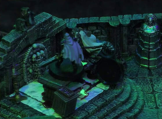 A battlemap of a stone crypt focused on Teor standing next to a wooden table on which an undead Raimond Davinos is standing. Talcydimir lays on the ground next to the table. The table sits on a raised part of the floor in front of a wall decorated with carved skull motifs. On a box in front of the table sits a tray bearing multiple surgical instruments. A silvery metal iron maiden is against a nearby wall. The room is lit by eerie green light.