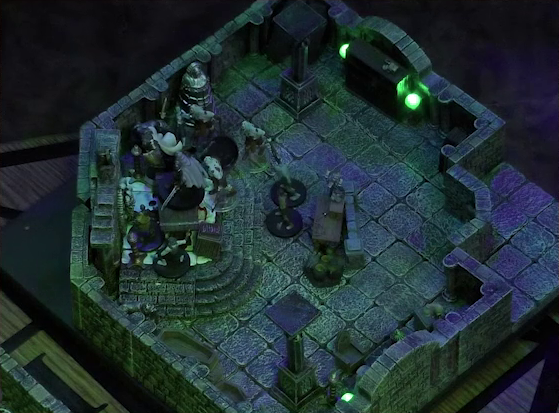 A battlemap of a stone crypt. Teor stands next to a wooden table on which an undead Raimond Davinos is standing. Talcydimir lays on the ground next to the table. A hoard of eight zombies begins to surround the table. The table sits on a raised part of the floor in front of a wall decorated with carved skull motifs. On a box in front of the table sits a tray bearing multiple surgical instruments. The room is lit by eerie glowing green torches.