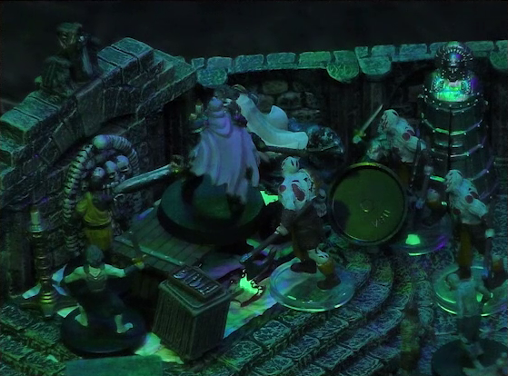 A battlemap of a stone crypt focused on Teor standing next to a wooden table on which an undead Raimond Davinos is standing. Talcydimir lays on the ground next to the table. A hoard of eight zombies surrounds the table. The table sits on a raised part of the floor in front of a wall decorated with carved skull motifs. On a box in front of the table sits a tray bearing multiple surgical instruments. A silvery metal iron maiden is against a nearby wall. The room is lit by eerie green light.