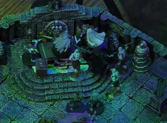 A battlemap of a stone crypt focused on Teor standing next to a wooden table on which an undead Raimond Davinos is standing. Talcydimir lays on the ground next to the table. A hoard of eight zombies surrounds the table. The table sits on a raised part of the floor in front of a wall decorated with carved skull motifs. On a box in front of the table sits a tray bearing multiple surgical instruments. A silvery metal iron maiden is against a nearby wall. The room is lit by eerie green light.