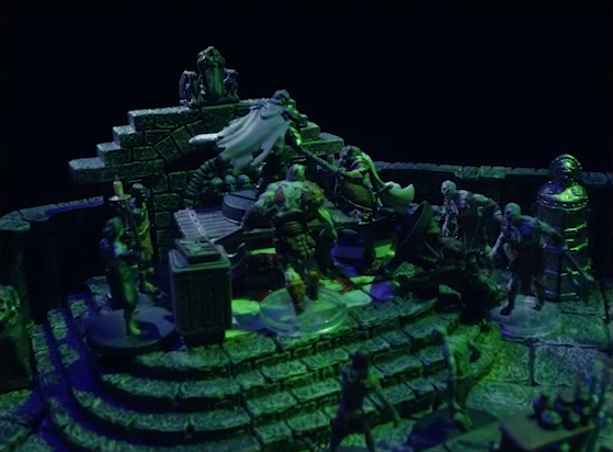 A dramatic shot of a battlemap of a stone crypt focused on Teor standing next to a wooden table on which an undead Raimond Davinos is standing. Talcydimir lays on the ground next to the table. A hoard of eight zombies surrounds the table. The table sits on a raised part of the floor in front of a wall decorated with carved skull motifs. On a box in front of the table sits a tray bearing multiple surgical instruments. A silvery metal iron maiden is against a nearby wall. The room is lit by eerie green light.