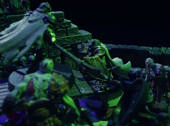 A dramatic shot of a battlemap of a stone crypt focused on Teor standing next to a wooden table on which an undead Raimond Davinos is standing. Talcydimir lays on the ground next to the table. A hoard of zombies surrounds the table. The room is lit by eerie green light.