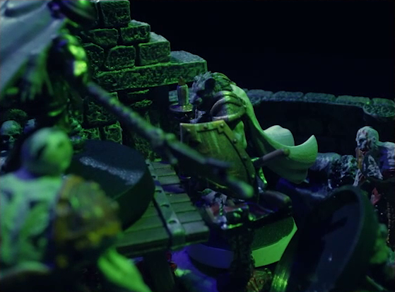 A dramatic shot of a battlemap of a stone crypt focused on Teor standing next to a wooden table on which an undead Raimond Davinos is standing. Teor is in focus in the photo, with much around him blurred. Talcydimir lays on the ground next to the table. A hoard of zombies surrounds the table. The room is lit by eerie green light.
