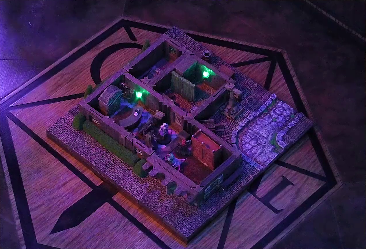 A battlemap of a street-side tailor shop eerily tinted purple and lit by glowing green torches. Kattigan, Wulferic, Tyranny, Bolaire, and Calastro Mercanaud stand in a main entry room in front of a curved front desk. A wardrobe is behind the desk and another one painted red is on the other side of the room. A door leads from the room to an adjoining one with curtains dividing it partially down the center. A third room is off to the side without a clear connection to the rest of the building. This room has stairs leading below ground. Outside the building is a wheelbarrow.